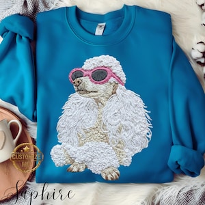 May include: Teal blue sweatshirt with an embroidered white poodle wearing pink sunglasses. The poodle is the main design, with detailed stitching. The word "Saphire" is written in cursive at the bottom.