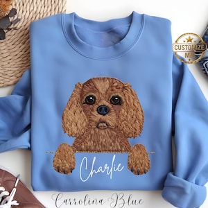 Puede incluir: Sudadera azul Carolina con un diseño bordado de perro Cavalier King Charles Spaniel. El nombre del perro, "Charlie", está escrito en cursiva debajo de la imagen. La sudadera tiene cuello redondo y mangas largas.
