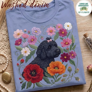 Op de afbeelding: Een lichtblauw t-shirt met een grafische print van een zwarte hond met krullend haar en een klein roze strikje op het hoofd, omringd door kleurrijke bloemen waaronder rode klaprozen, oranje klaprozen en diverse roze en witte cosmosbloemen.