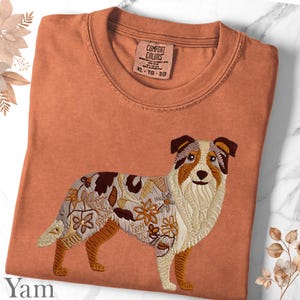 May include: A terracotta-coloured t-shirt with an embroidered dog design and floral accents. The dog has brown, white, and tan fur. The shirt has a crew neck and a Comfort Colors tag. The size is XL-TG-EG. The word "Yam" is at the bottom left.