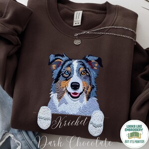 Könnte beinhalten: Dunkelbraunes Sweatshirt mit einem gestickten Australian Shepherd Hund mit blauem, braunem und weißem Fell. Der Name "Kriket" ist darunter gestickt. Die Worte "Dark Chocolate" sind unten aufgedruckt.
