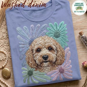 May include: A light blue t-shirt featuring a printed graphic of a curly-haired brown dog surrounded by light blue, teal, and light purple daisy-like flowers. The text "Washed denim" and a circular badge stating "LOOKS LIKE EMBROIDERY BUT IT'S PRINTED!" are visible.