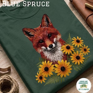 Puede incluir: Una camiseta verde oscuro con un gráfico de un zorro rojo rodeado de flores amarillas y hojas de helecho verdes. El texto "BLUE SPRUCE" está impreso en blanco en la parte superior. El diseño incluye texturas de pelaje y elementos florales.