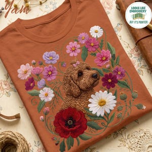 Può includere: Una t-shirt color ruggine con una stampa grafica di un cane marrone dal pelo riccio con un piccolo fiocco rosa sulla testa, circondato da una colorata composizione di fiori rosa, viola, bianchi e rossi con steli e foglie verdi.