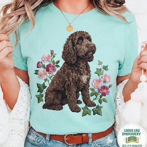 May include: A light blue t-shirt with a brown dog illustration surrounded by pink and purple flowers and green leaves. The text "Chalky Mint" is at the bottom of the shirt. The shirt also has the text "Looks like embroidery but it's printed!"