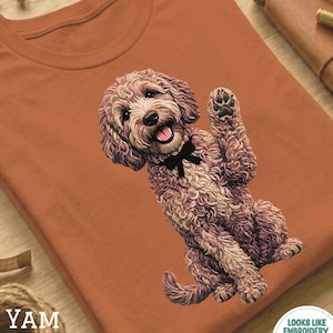 Puede incluir: Una camiseta de color óxido con una ilustración detallada de un perro sonriente de pelo rizado con una pajarita negra. El perro está saludando con la pata. El texto "YAM" está impreso en la camiseta.