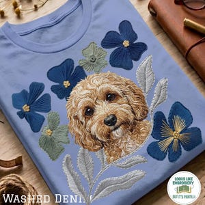 Può includere: Una t-shirt azzurro chiaro con una stampa raffigurante un cane dal pelo riccio color marrone dorato, circondato da illustrazioni in stile ricamo di fiori blu scuro e verde salvia con foglie grigio argento. Il testo recita: LOOKS LIKE EMBROIDERY BUT IT'S PRINTED.
