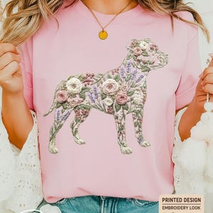 May include: A light pink t-shirt featuring a floral dog design. The dog is composed of embroidered-style flowers in shades of pink, white, and purple. The word "Blossom" is written in cursive at the bottom of the shirt.