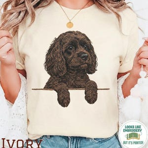 May include: Ivory-colored t-shirt featuring a detailed illustration of a brown dog with curly fur. The dog's paws rest on a horizontal line. The word "IVORY" is printed at the bottom. A circular sticker reads "LOOKS LIKE EMBROIDERY BUT IT'S PRINTED!"