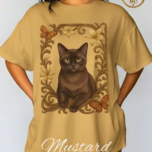 Könnte beinhalten: Senffarbenes T-Shirt mit einer braunen Katze in einem floralen Rahmen mit Schmetterlingen. Das Wort "Mustard" ist unten aufgedruckt. Das Shirt hat kurze Ärmel und eine lockere Passform.