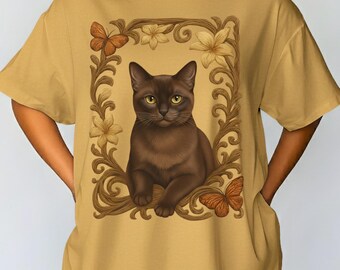 Può includere: T-shirt color senape con un gatto marrone all'interno di una cornice floreale con farfalle. La parola "Mustard" è stampata in basso. La maglietta ha maniche corte e una vestibilità ampia.