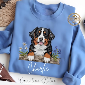 May include: A Carolina Blue sweatshirt with a Bernese Mountain Dog design. The dog is depicted in black, brown, and white, with a happy expression. The name "Charlie" is written below the dog, with a gold seal that says "Customize Me!"