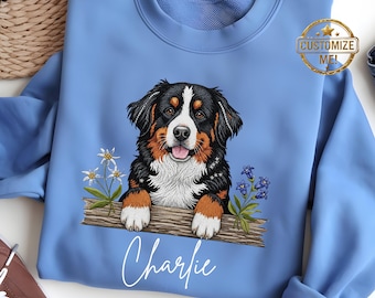 Puede incluir: Sudadera azul Carolina con un diseño de perro de montaña bernés. El perro es negro, marrón y blanco, con una expresión sonriente. El nombre "Charlie" está escrito debajo, con un sello dorado "Customize Me!"