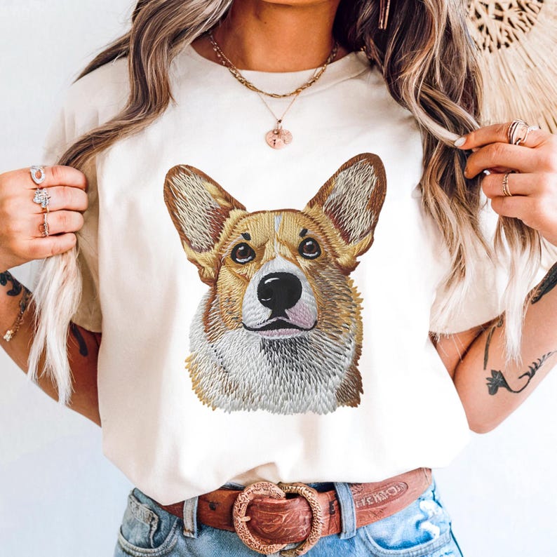 K&ouml;nnte beinhalten: Cremefarbenes T-Shirt mit einem detaillierten, gestickten Portr&auml;t eines Corgi-Hundes. Der Hund hat braunes und wei&szlig;es Fell, gro&szlig;e Ohren und eine schwarze Nase. Das Shirt wird mit einer Jeans und einem braunen G&uuml;rtel getragen.
