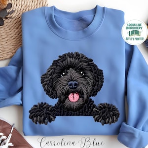 May include: Carolina Blue sweatshirt featuring a black dog graphic with a pink tongue. The dog appears to be embroidered. The text "Looks like embroidery but it's printed!" is in a circle on the upper right. The text "Carrolina Blue" is at the bottom.