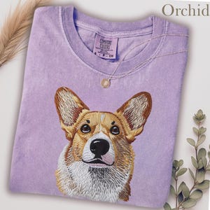 K&ouml;nnte beinhalten: Orchideenfarbenes lila T-Shirt mit einem gestickten Corgi-Hunde-Gesicht. Das Shirt hat einen Rundhalsausschnitt und kurze &Auml;rmel. Der Markenname "Comfort Colors" ist sichtbar. Eine goldene Halskette mit Anh&auml;nger liegt auf dem Shirt.