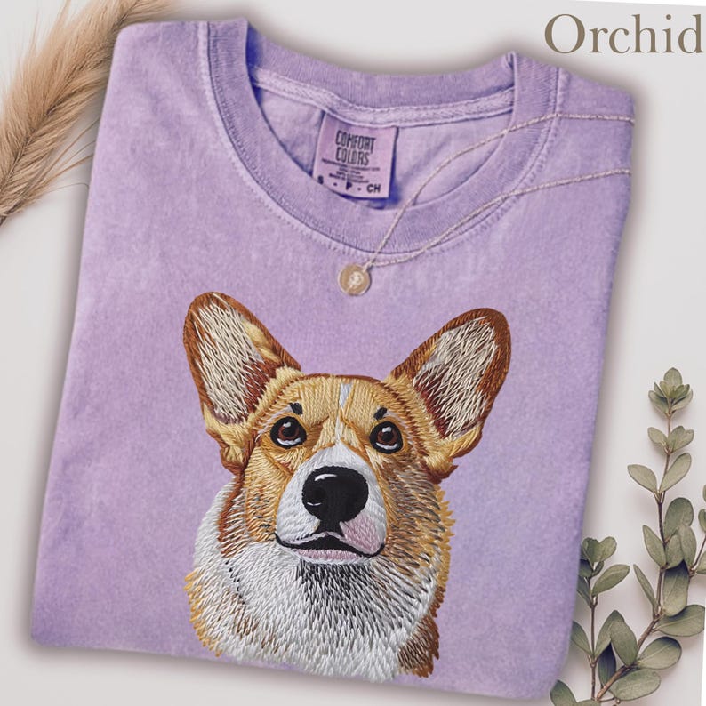 K&ouml;nnte beinhalten: Orchideenfarbenes lila T-Shirt mit einem gestickten Corgi-Hunde-Gesicht. Das Shirt hat einen Rundhalsausschnitt und kurze &Auml;rmel. Der Markenname "Comfort Colors" ist sichtbar. Eine goldene Halskette mit Anh&auml;nger liegt auf dem Shirt.