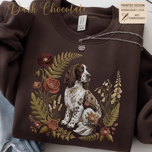 Könnte beinhalten: Dunkelbraunes Sweatshirt mit einem aufgedruckten Design in Stickoptik. Das Design zeigt einen braunen und weißen Hund, umgeben von floralen Elementen in Rot-, Grün- und Brauntönen. Der Text "Dark Chocolate" befindet sich oben links.