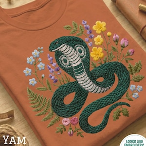 Op de afbeelding: Roestkleurig T-shirt met een gedetailleerd geborduurd ontwerp van een groene cobra met witte accenten, omringd door kleurrijke bloemen en bladeren. De tekst "YAM" is zichtbaar aan de linkerkant. Het shirt heeft een ronde hals en korte mouwen.