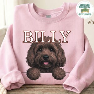 May include: A light pink crewneck sweatshirt featuring a graphic of a brown dog with curly fur and the name BILLY printed in large brown letters above it. A circular badge in the top right corner reads LOOKS LIKE EMBROIDERY BUT IT'S PRINTED.