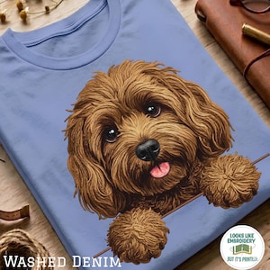 Peut inclure: Un t-shirt bleu clair avec une illustration graphique d'un chien brun aux poils bouclés, aux grands yeux sombres et à la langue rose tirée. Le chien semble regarder par-dessus une ligne horizontale. Le texte "WASHED DENIM" est imprimé en bas.