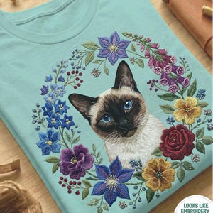 Könnte beinhalten: Hellblaues T-Shirt mit einem Siamkatzen-Motiv, umgeben von einem Blumenkranz. Der Kranz enthält blaue, violette, gelbe und rote Blumen. Der Text "CHALKY MINT" befindet sich am unteren Rand des T-Shirts.