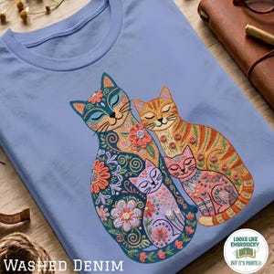 Può includere: Una t-shirt azzurra con un disegno stampato di tre gatti con intricati motivi floreali e a spirale. Il gatto più grande è blu, quello centrale è arancione a strisce e il più piccolo è rosa. Tutti i gatti hanno gli occhi chiusi.