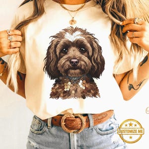 May include: Cream-colored t-shirt featuring a detailed illustration of a brown and white dog. The dog has a decorative collar. The shirt is worn with blue denim shorts and a brown belt with a gold-colored buckle. The text "CUSTOMIZE ME" is on the bottom right.
