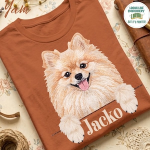 May include: A rust-colored t-shirt featuring a graphic of a fluffy, light-brown Pomeranian dog with its paws resting on a horizontal line. Below the dog, the name Jacko is printed in white serif font. A circular badge reads Looks like embroidery but it's printed.
