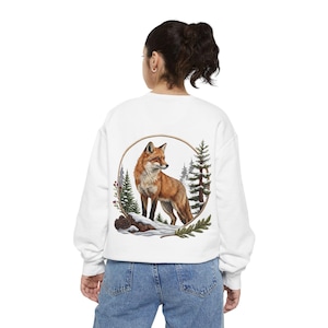 May include: A white long-sleeve sweatshirt featuring a circular graphic on the back. The design depicts a red fox standing on a snow-covered log, surrounded by evergreen trees and a thin brown circular frame against a white background.
