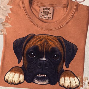 May include: A terracotta-colored t-shirt featuring a detailed illustration of a Boxer dog's face. The dog has brown and black fur, with a white patch on its nose and paws. The shirt has a crew neck and a tag that reads "Comfort Colors". The word "Yam" is written at the bottom.
