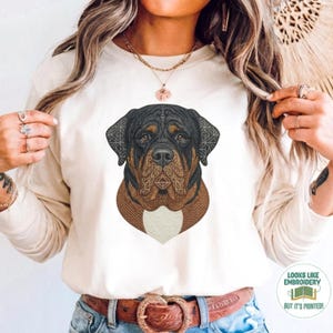 May include: Cream-colored long-sleeve shirt featuring a detailed embroidered-style Rottweiler dog portrait. The dog's head is black and brown with intricate patterns. The shirt has a white bib-like shape at the bottom of the dog's chest.