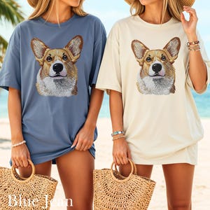 K&ouml;nnte beinhalten: Zwei &uuml;bergro&szlig;e T-Shirts, eines in Blau und eines in Creme, jeweils mit einem detaillierten, gestickten Corgi-Gesicht. Die Shirts werden mit Stroh-Tragetaschen getragen. Der Text "Blue Jean" ist unten im Bild zu sehen.