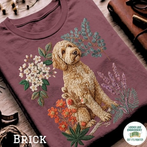 Op de afbeelding: Baksteenkleurig T-shirt met een geborduurd ontwerp van een golden doodle hond omringd door bloemen en bladeren. Het ontwerp bevat witte, oranje en paarse bloemelementen. Het woord "BRICK" staat onderaan gedrukt.