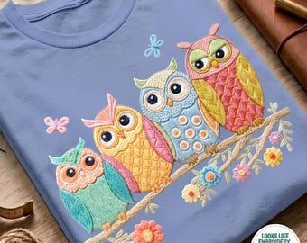 May include: A light blue t-shirt featuring an embroidered design of four colorful owls perched on a branch. The owls are teal, yellow, light blue, and pink with textured patterns. Small embroidered flowers and butterflies surround the owls.