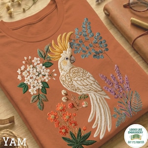 May include: Terracotta-coloured t-shirt with an embroidered design of a white cockatoo with a yellow crest, surrounded by flowers and foliage. The design includes white, orange, purple, and blue elements. The text "YAM" is visible at the bottom left.