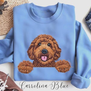 May include: A Carolina Blue sweatshirt with an embroidered brown dog design. The dog has its tongue out and its paws are at the bottom of the sweatshirt. The text "Carolina Blue" is written in cursive at the bottom.