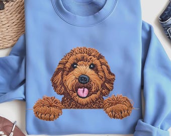 May include: Carolina Blue sweatshirt featuring an embroidered brown dog with a pink tongue. The dog's paws appear to be holding the bottom of the sweatshirt. The text "Carolina Blue" is written in cursive at the bottom of the sweatshirt.