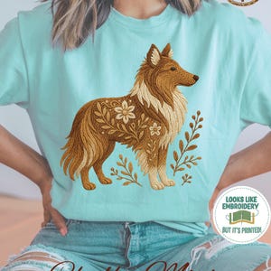 May include: A light mint green t-shirt featuring a graphic of a brown collie dog with floral and leaf patterns integrated into its fur. The shirt includes a circular badge in the top right corner that says CUSTOMIZE ME! and a bottom right badge that says LOOKS LIKE EMBROIDERY BUT IT'S PRINTED!