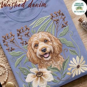 Könnte beinhalten: Ein hellblaues T-Shirt mit einem Druckmotiv eines goldbraunen, lockigen Hundes, umgeben von grünen Blättern, braunen Zweigen und weißen Blumen. Oben steht der Text "Washed denim" und ein rundes Abzeichen besagt "LOOKS LIKE EMBROIDERY BUT IT'S PRINTED!"