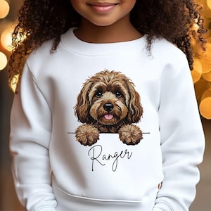 May include: White sweatshirt with a brown dog illustration and the name "Ranger" in cursive. The dog has brown fur and is peeking over a white horizontal line. The sweatshirt is a plain white colour.
