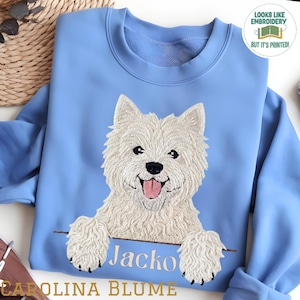 May include: A light blue crewneck sweatshirt featuring a printed graphic of a white West Highland White Terrier dog with its paws resting on a horizontal line above the name Jacko. A circular badge in the corner reads Looks Like Embroidery But It's Printed.