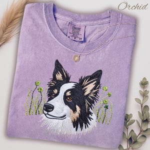 Może przedstawiać: Lawendowa koszulka Comfort Colors z haftowanym portretem psa Border Collie i zielonymi detalami kwiatowymi. Złożona koszulka ma okrągły dekolt. Złoty naszyjnik z wisiorkiem jest zarzucony na koszulkę. Słowo "Orchid" jest napisane kursywą.