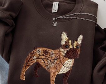 May include: A dark brown sweatshirt with an embroidered French Bulldog design. The dog is detailed with brown, tan, and cream thread, featuring floral accents. A silver necklace with a small pendant rests on the folded garment. The text "Dark Chocolate" is written in cursive.