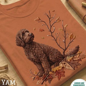 May include: A terracotta-coloured t-shirt with a detailed illustration of a brown dog surrounded by autumn leaves and branches. The design includes the text "YAM" on the lower left and "LOOKS LIKE EMBROIDERY BUT IT'S PRINTED!" on the lower right.