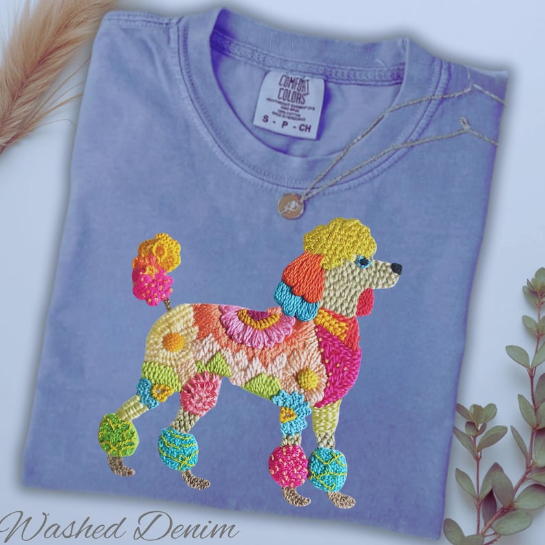 Pu&ograve; includere: T-shirt azzurra con un design di barboncino ricamato. Il barboncino &egrave; decorato con motivi floreali colorati nelle tonalit&agrave; del rosa, arancione, giallo e verde. Il testo "Washed Denim" &egrave; in basso.