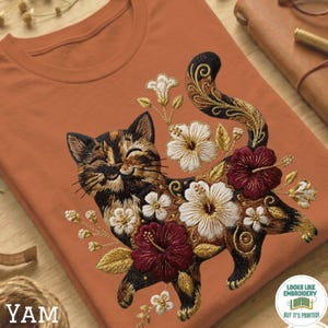 以下が含まれることがあります： バーントオレンジ色のTシャツ。サビ猫の刺繍デザインが施されており、白と濃い赤のハイビスカスの花、金色の葉、小さな蕾に囲まれています。猫は目を閉じて微笑んでいるような表情をしており、ひげも刺繍されています。