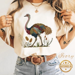 Puede incluir: Camiseta color marfil con una ilustración detallada de un emú con plumas coloridas y cuello largo, de pie entre plantas estilizadas. También se ve el texto "Ivory" y "Customize Me".