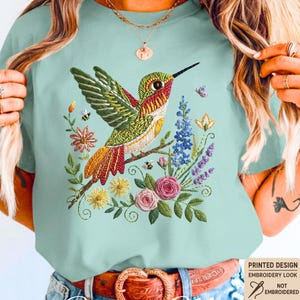 Op de afbeelding: Lichtblauw T-shirt met een geborduurd ontwerp van een kolibrie op een tak met kleurrijke bloemen en bijen. De tekst "Chalky Mint" staat onderaan het shirt.