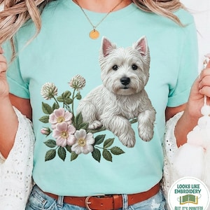 Può includere: T-shirt color menta con un'illustrazione di un cane West Highland White Terrier, fiori rosa e foglie verdi. Il testo "LOOKS LIKE EMBROIDERY BUT IT'S PRINTED!" è stampato sulla maglietta.